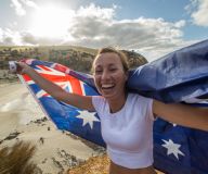 Kvinna med australiska flaggan vid kusten, ler mot kameran under en solig dag.