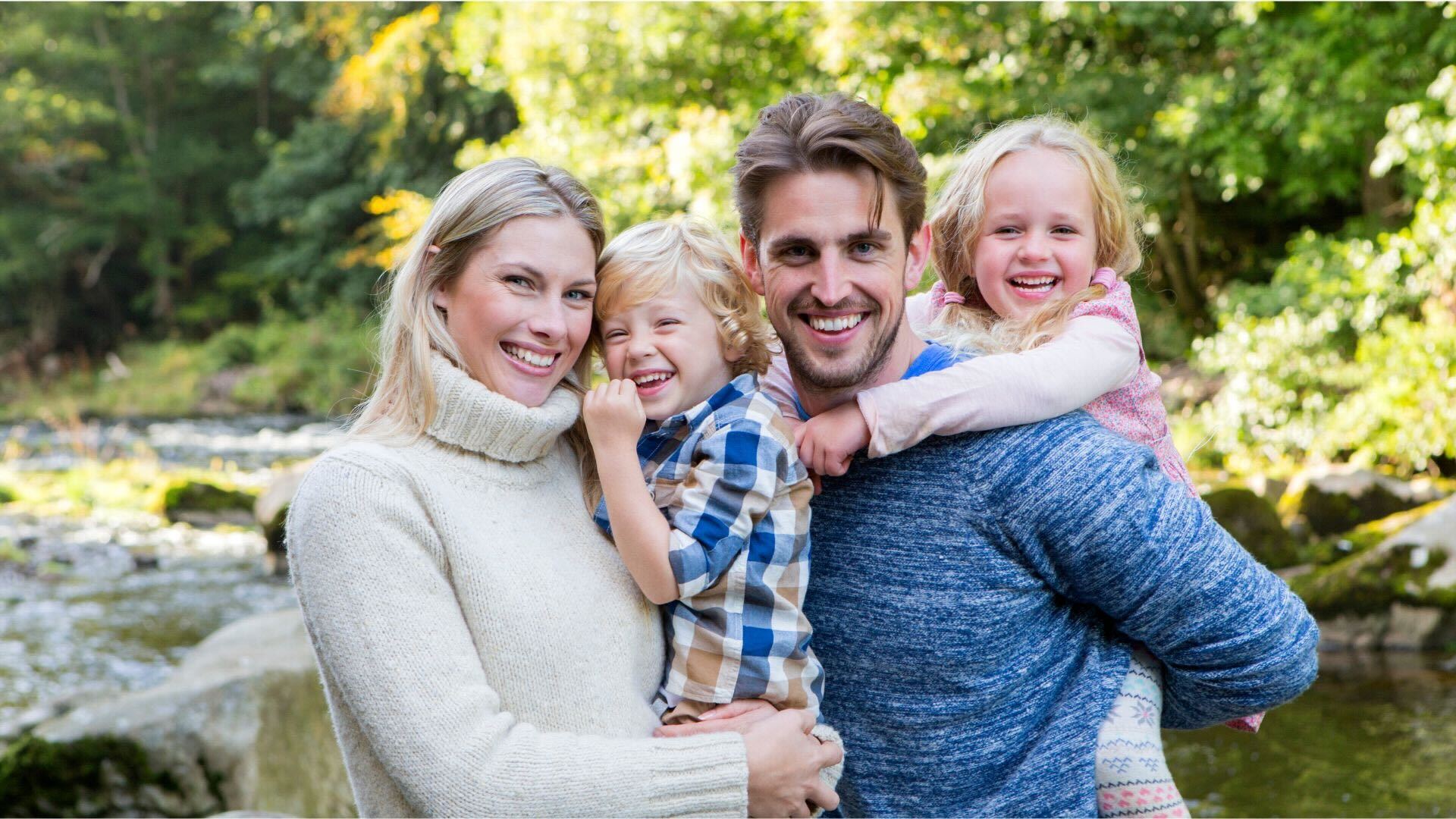 Familj med två barn vid en bäck, glada och leende i naturskön miljö.